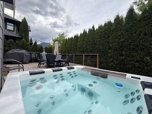 A hot tub with jets, surrounded by a deck with chairs and a table, set against a backdrop of dense trees and a cloudy sky. - Luxury Mountain Retreat in Squamish (Squamish)