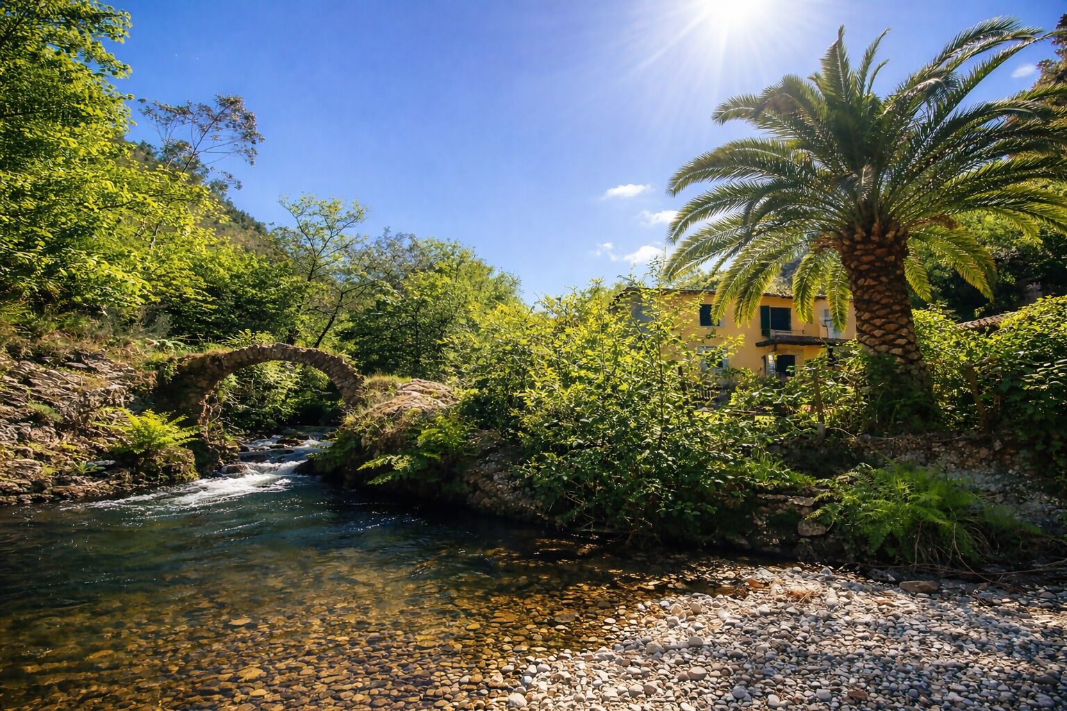 Un río con un puente de piedra, abundante vegetación y un edificio amarillo con una palmera.