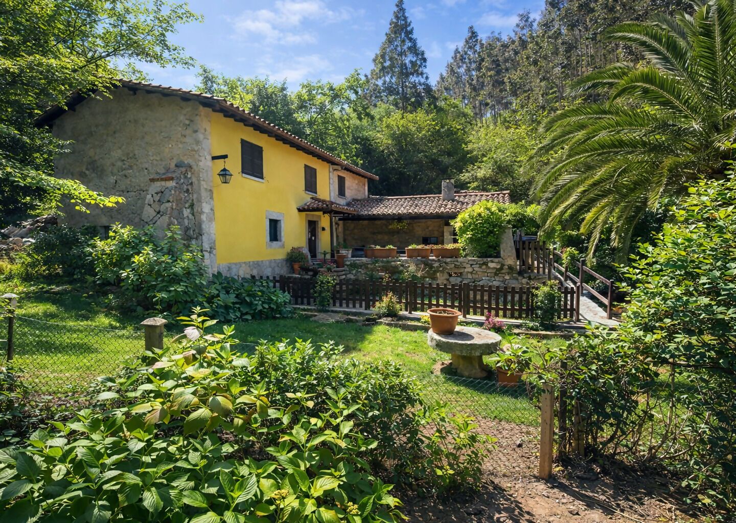 Un bâtiment rustique de couleur jaune, doté d’une façade en pierre, entouré d’une végétation luxuriante et d’une clôture en bois.