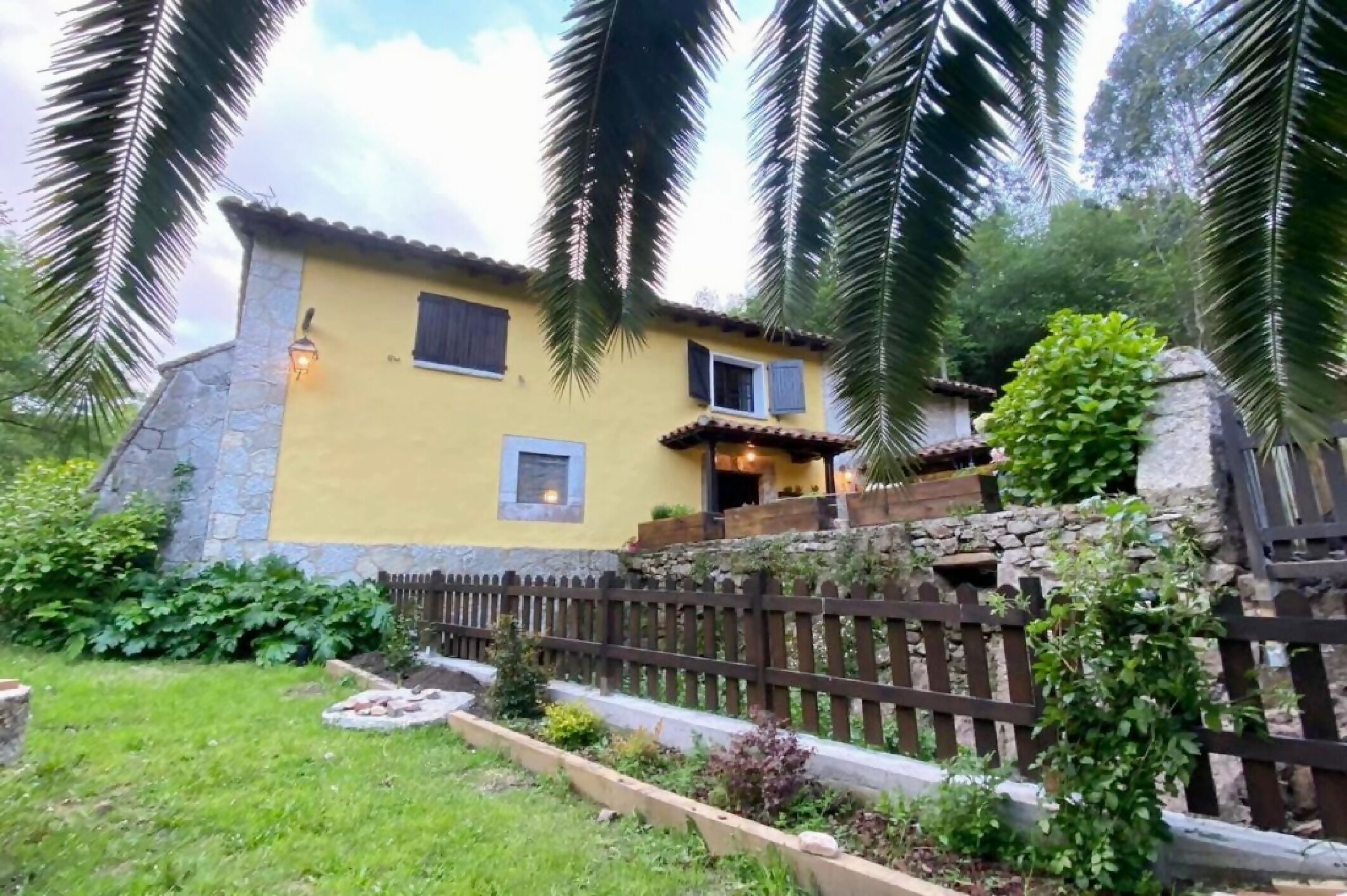 A yellow building with a stone base, a wooden fence, and a garden with various plants.