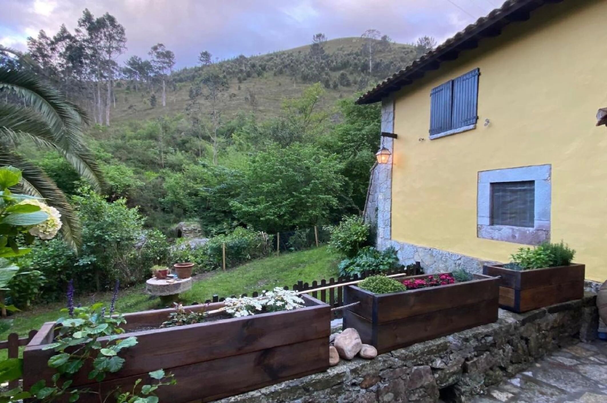 A yellow building with blue shutters, surrounded by a garden with wooden planters and a stone wall.