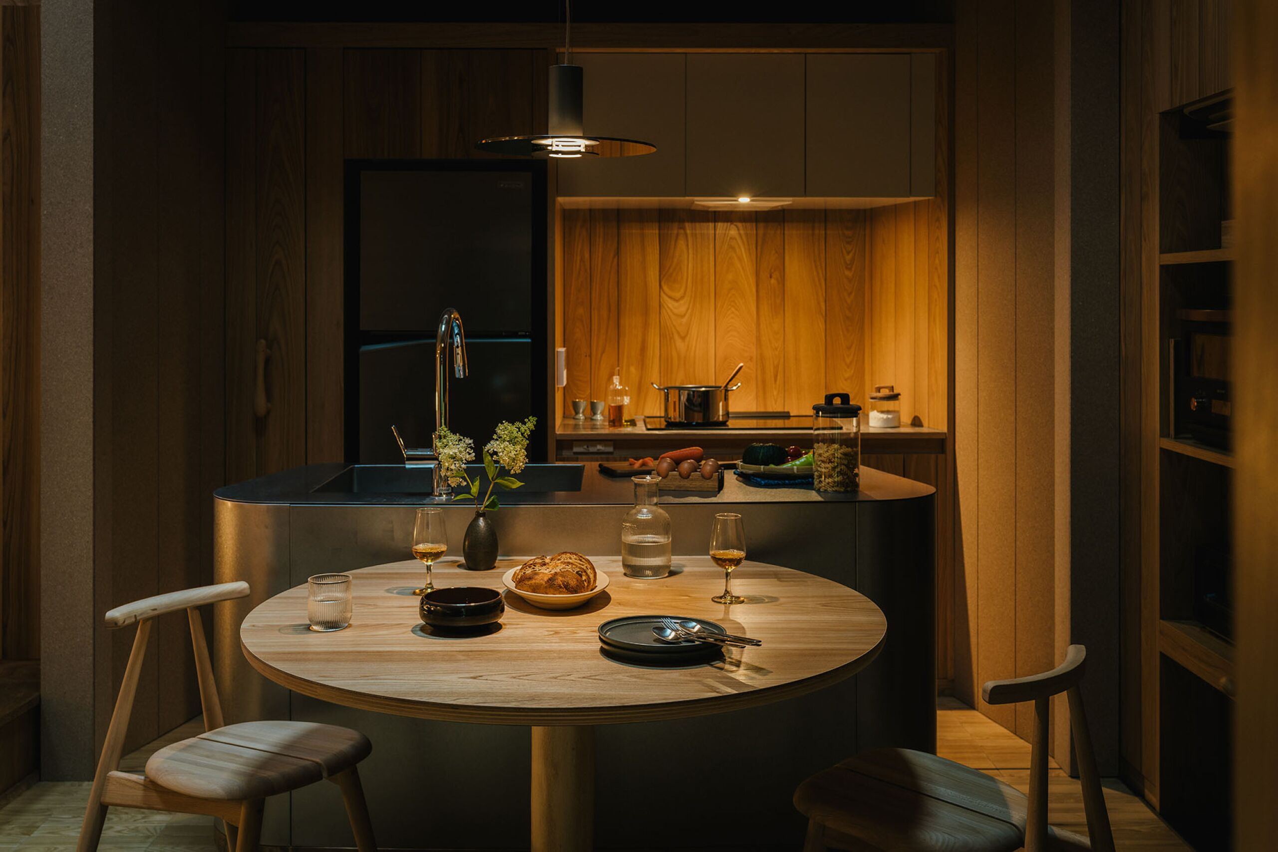 A modern kitchen with wooden cabinets, a central island with a round table, and a hanging light fixture.