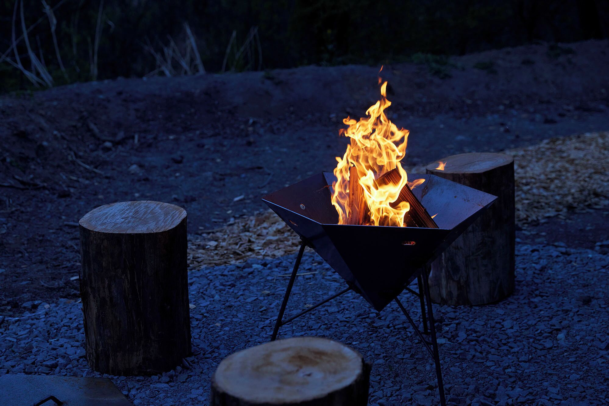 Un foyer métallique avec des flammes, entouré de trois troncs d’arbres en bois, le tout posé sur une surface de gravier.