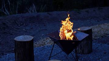 Un foyer métallique avec des flammes, entouré de trois troncs d’arbres en bois, le tout posé sur une surface de gravier.