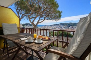 Een houten tafel met twee koffiekopjes en een glas sinaasappelsap, een bord croissants, en een stoel op een balkon met uitzicht over een stadsbeeld en een boom.