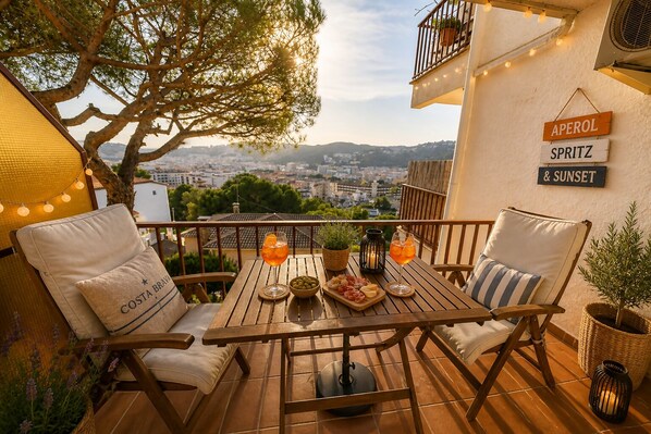 A wooden table with two chairs, a bottle of Aperol Spritz, a candle, and a small plant on an outdoor balcony with a city view.