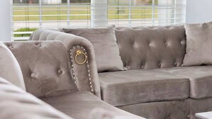 Un sofá gris con tapicería texturizada, adornado con detalles de anillos y cuentas, ubicado en una habitación con amplios ventanales y persianas.