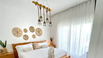 A modern bedroom with a wooden bed, a bedside table, a hanging light fixture, and a view of the city through sheer curtains.