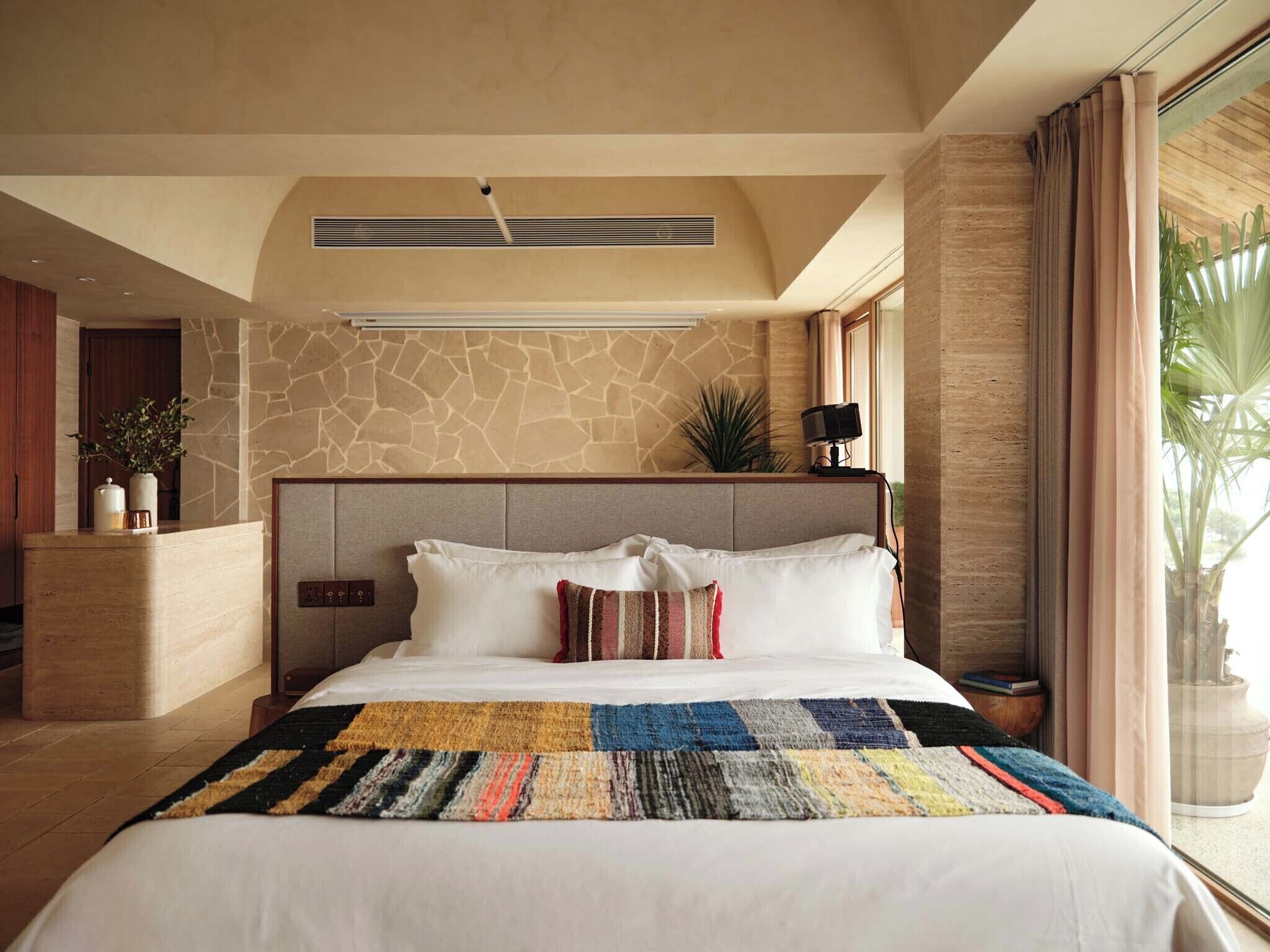 A bedroom with a large bed, a stone wall, a nightstand, a lamp, and a window with curtains.