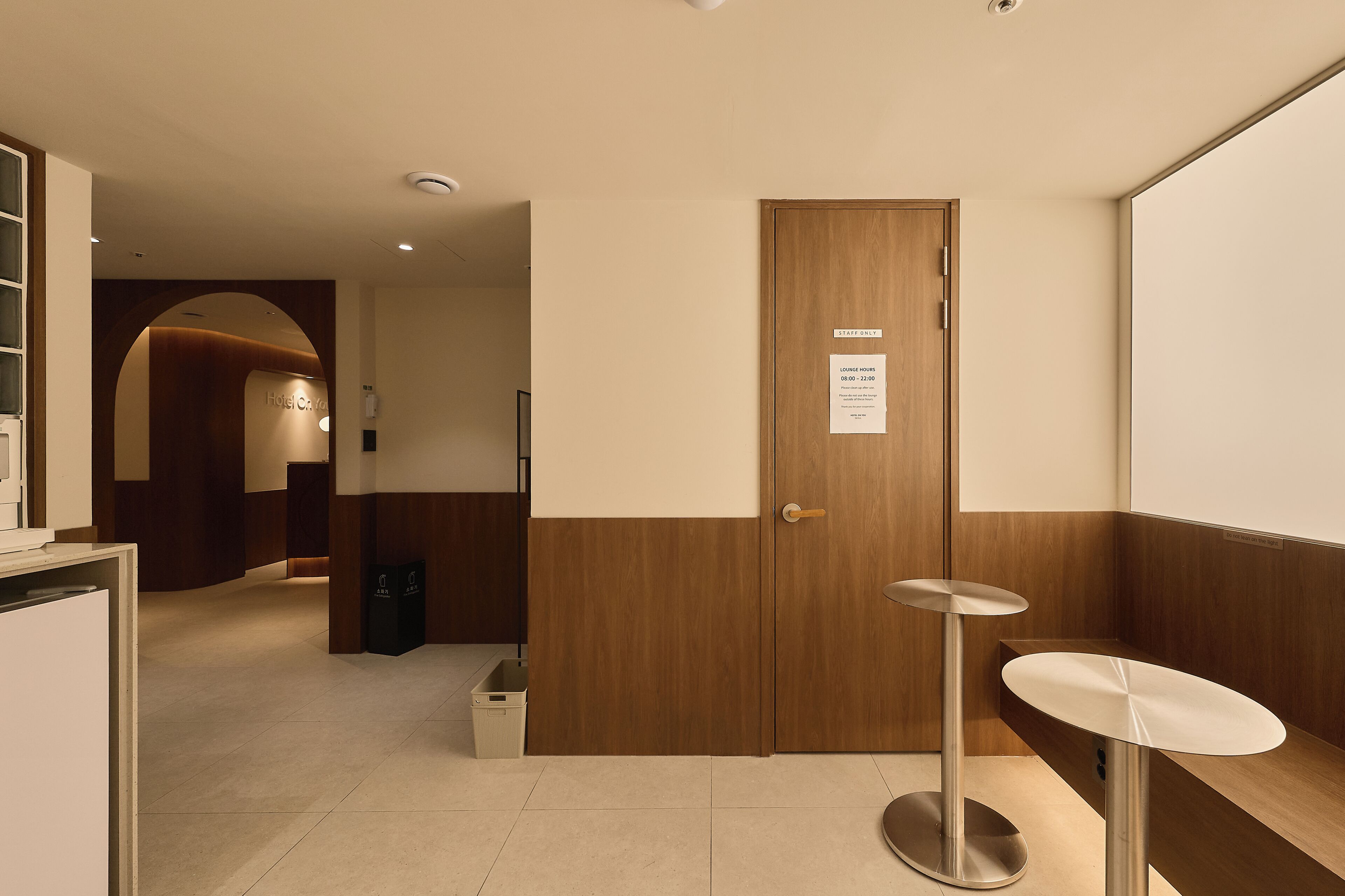Un vestíbulo de hotel con un mostrador de recepción, una puerta de madera con un cartel y un cubo de basura pequeño.