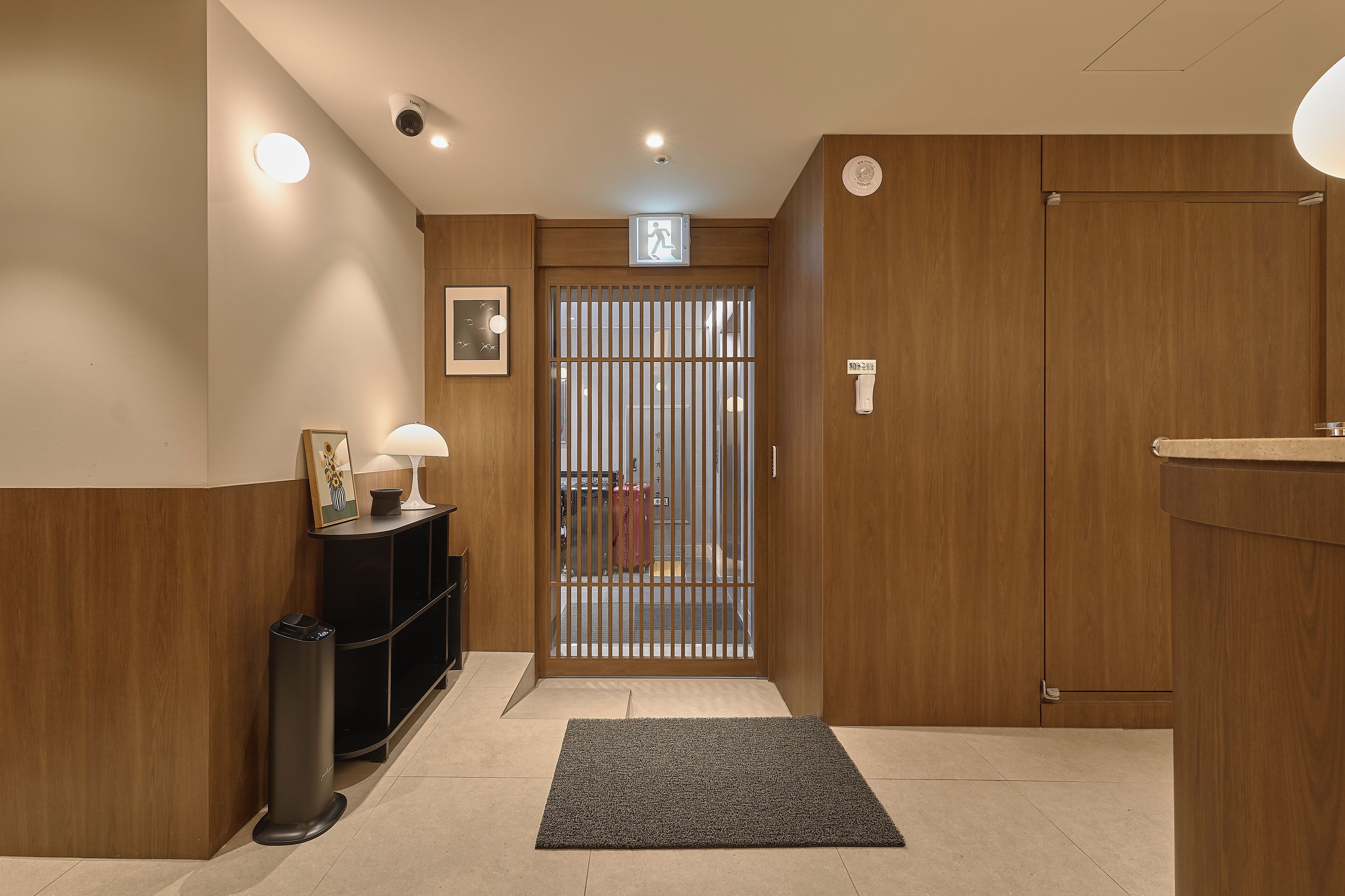 A hotel entrance with a wooden reception desk, a black trash bin, a grey mat, and a door with a metal gate.