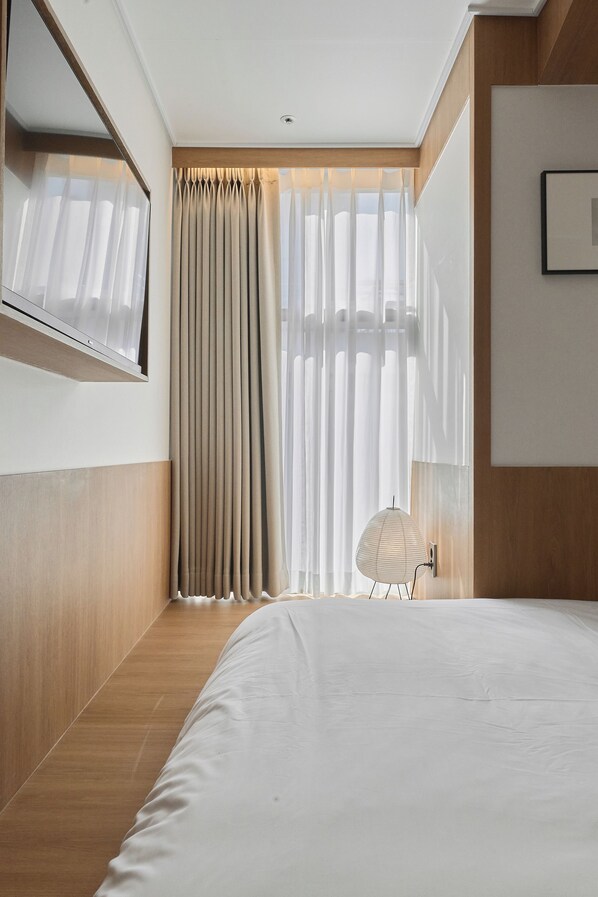 Ein modernes Hotelzimmer mit einem großen Bett, einem wandmontierten Spiegel, einem Fenster mit durchsichtigen Vorhängen und einem kleinen Tisch mit Lampe.