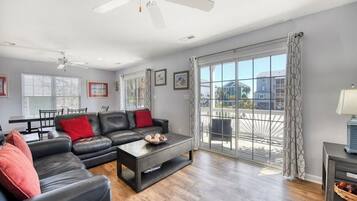 Un vaste salon comprenant un canapé en cuir noir, une table basse et un coin repas avec une table et des chaises. Un balcon offre une vue sur le quartier.