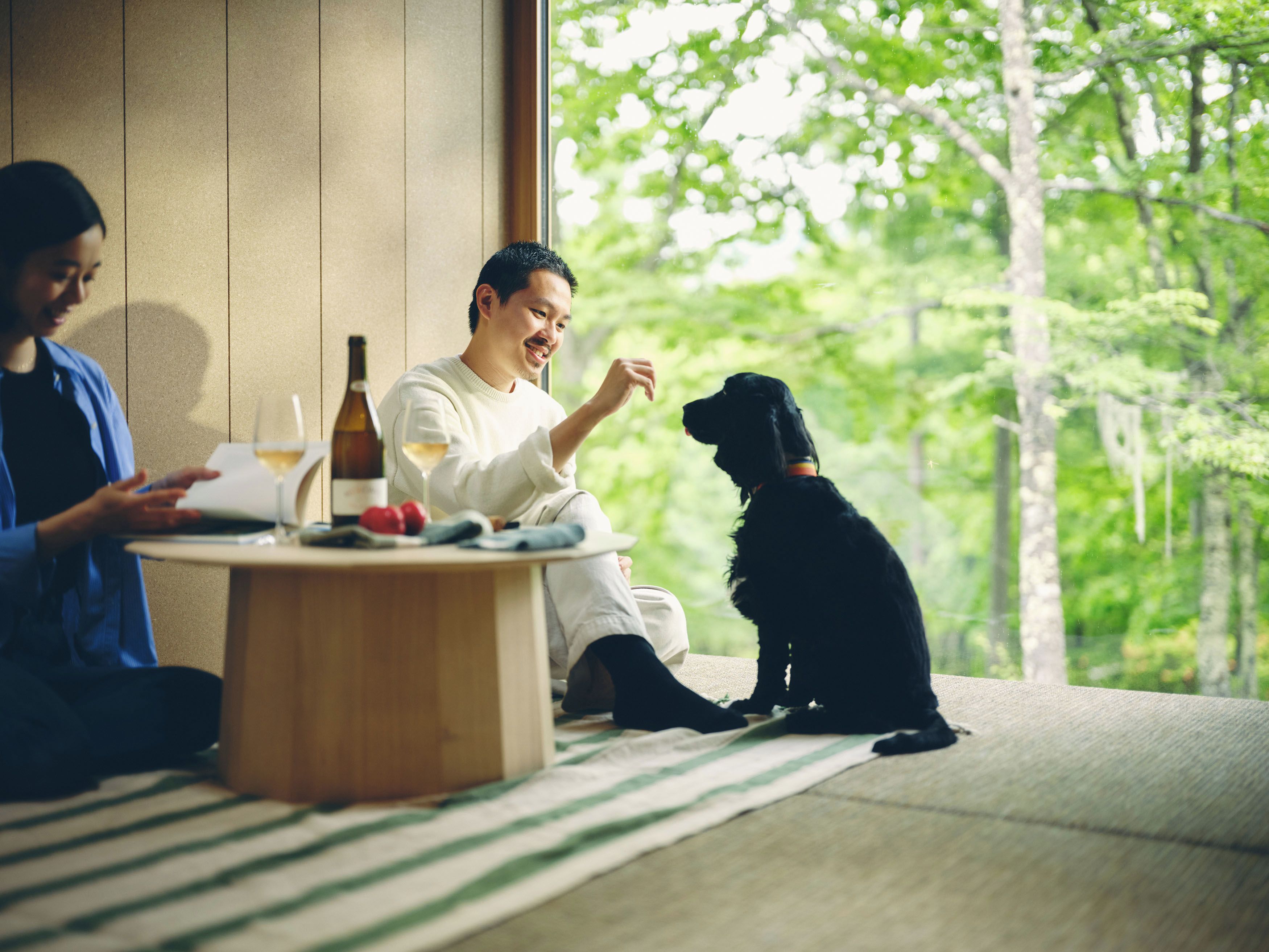 A room with a large window, a round wooden table, and a black dog sitting on a striped mat.