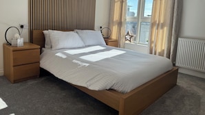 A neatly made bed with a headboard, a bedside table with a lamp, and a window with curtains. - 2 bedroom deluxe apartment in Leicester (Leicester)
