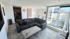A modern living room with a grey sectional sofa, a coffee table, and a large window offering a city view. - 2 bedroom deluxe apartment in Leicester (Leicester)