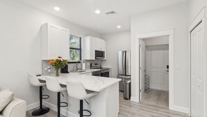 Una cocina moderna con armarios blancos, una isla central con taburetes de bar, un microondas, un refrigerador y un ventanal con persianas.