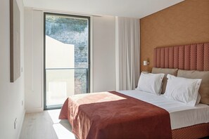 A modern hotel room with a large bed, a view of a stone wall, and a sliding glass door to an outdoor area. - Riverside Luxe 2BR | Pool, Spa, Balcony and Parking (Vila Nova de Gaia)