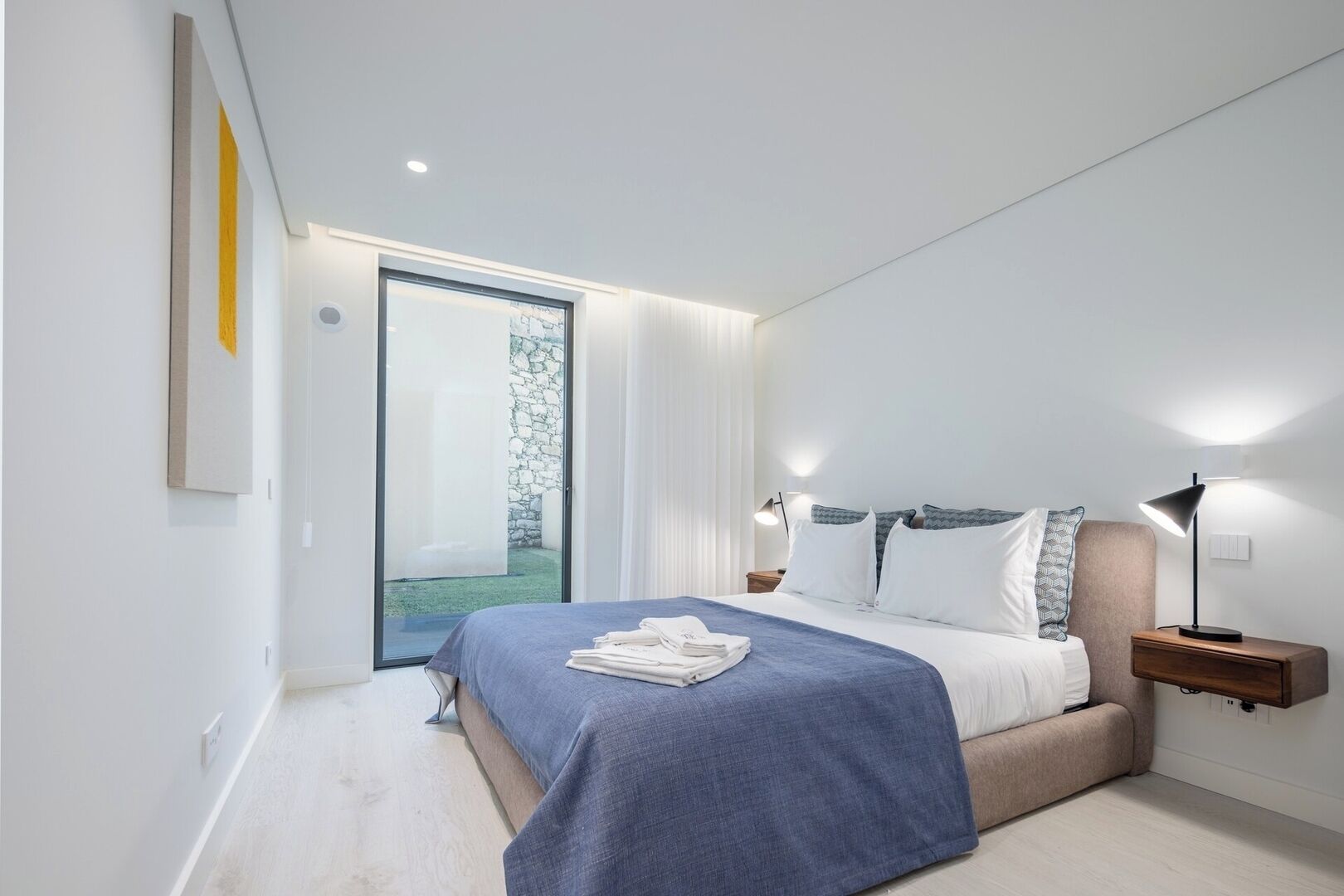 A modern bedroom with a bed, bedside tables, and a view of a garden through a glass door.