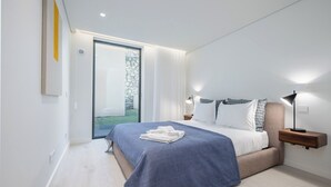 Quarto moderno com cama, mesas de cabeceira e vista para um jardim através de uma porta de vidro.