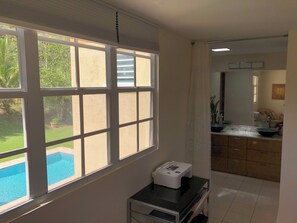 A room with a large window overlooking a swimming pool and greenery, a kitchen area with a sink and cabinets, and a printer on a small table. - A Private Paradise by the Rainforest in Sunny Luquillo (Pitahaya)