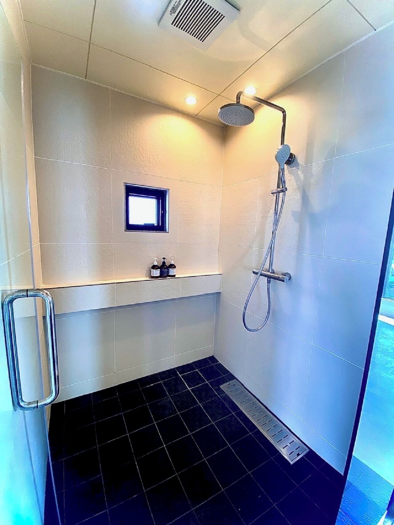 A small, modern bathroom with a shower, a window, and a shelf with toiletries.