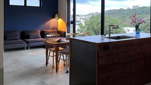 Una cocina moderna con una isla grande, un comedor con mesa y sillas, un sofá y vistas a un paisaje urbano a través de amplios ventanales.