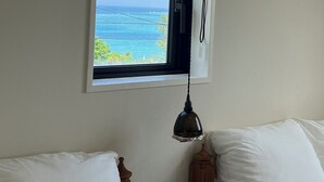 Un dormitorio con un ventanal que ofrece vistas al océano. Hay una cama con ropa de cama blanca y cabecera de madera.