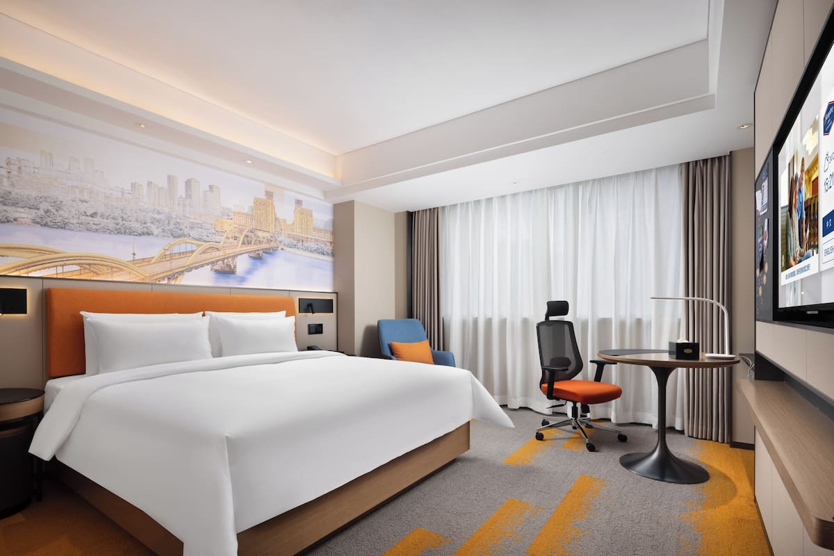 Ein Hotelzimmer mit einem großen Bett, einem Schreibtisch, einem Sessel, einem Fernseher und einer Wandmalerei mit Stadtansicht.