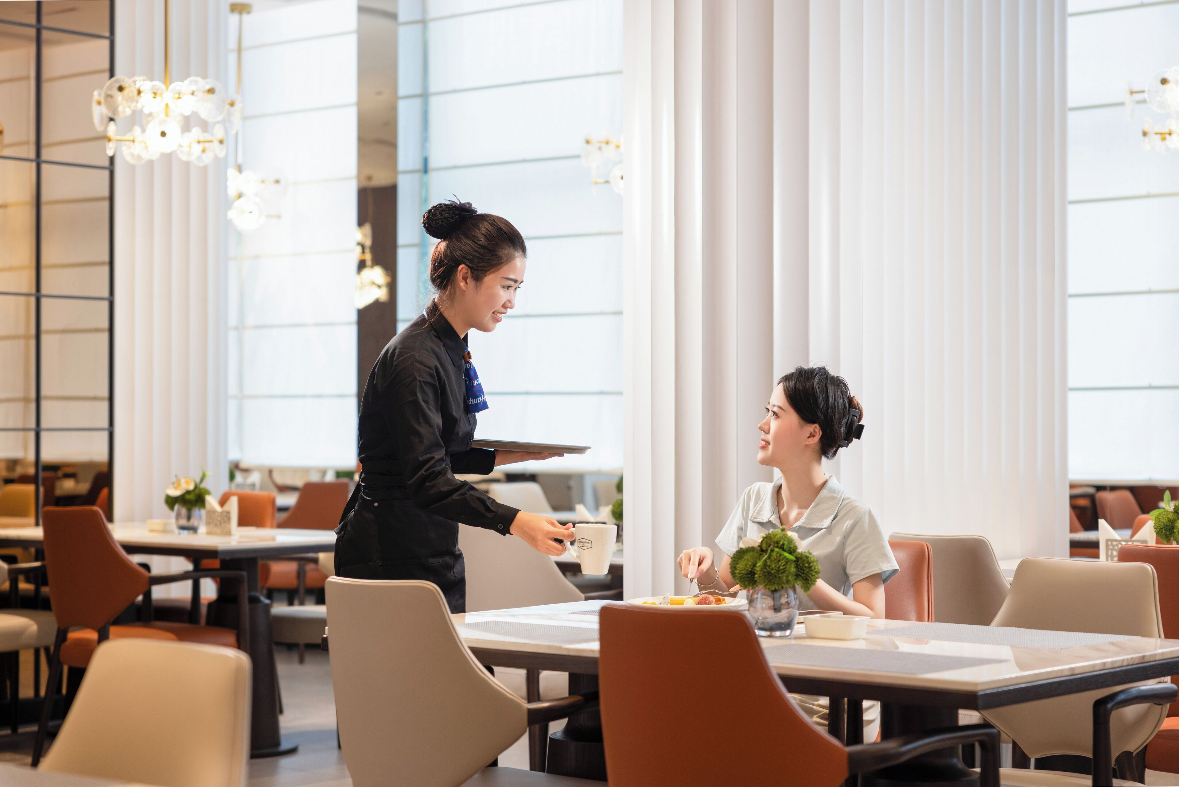 A modern dining area with tables and chairs, a waiter serving a customer, and large windows with vertical blinds.
