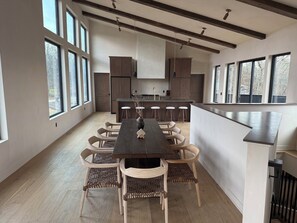 A spacious dining area with a long table and wicker chairs, a kitchen with wooden cabinets, and large windows providing natural light.