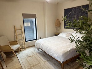 A bedroom with a bed, a chair, a ladder, a window, a wall art piece, and a hanging light fixture.