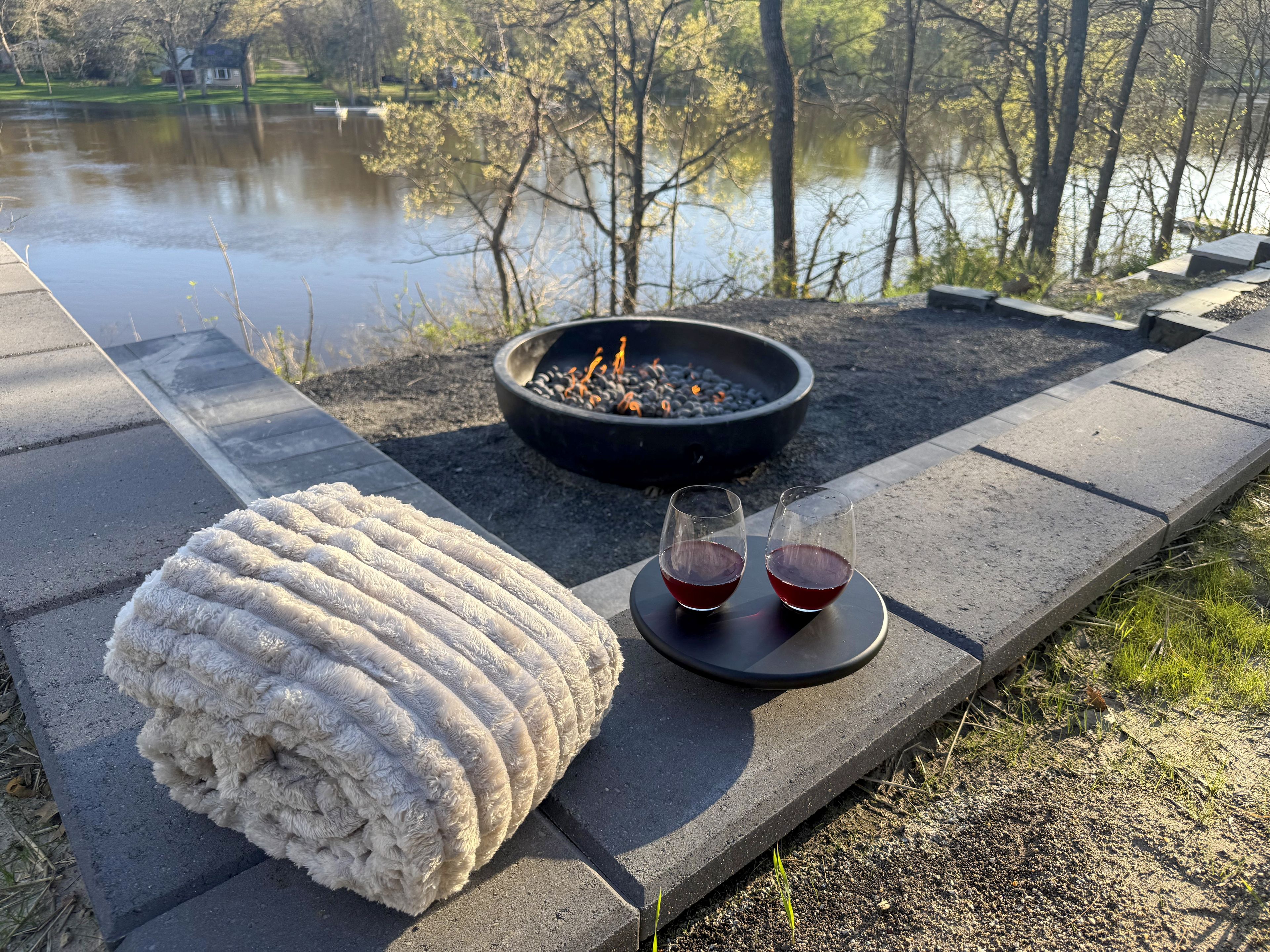 A fire pit with two glasses of red drink, a white rug, and a view of a lake.