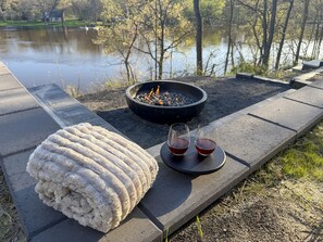 A fire pit with two glasses of red drink, a white rug, and a view of a lake.