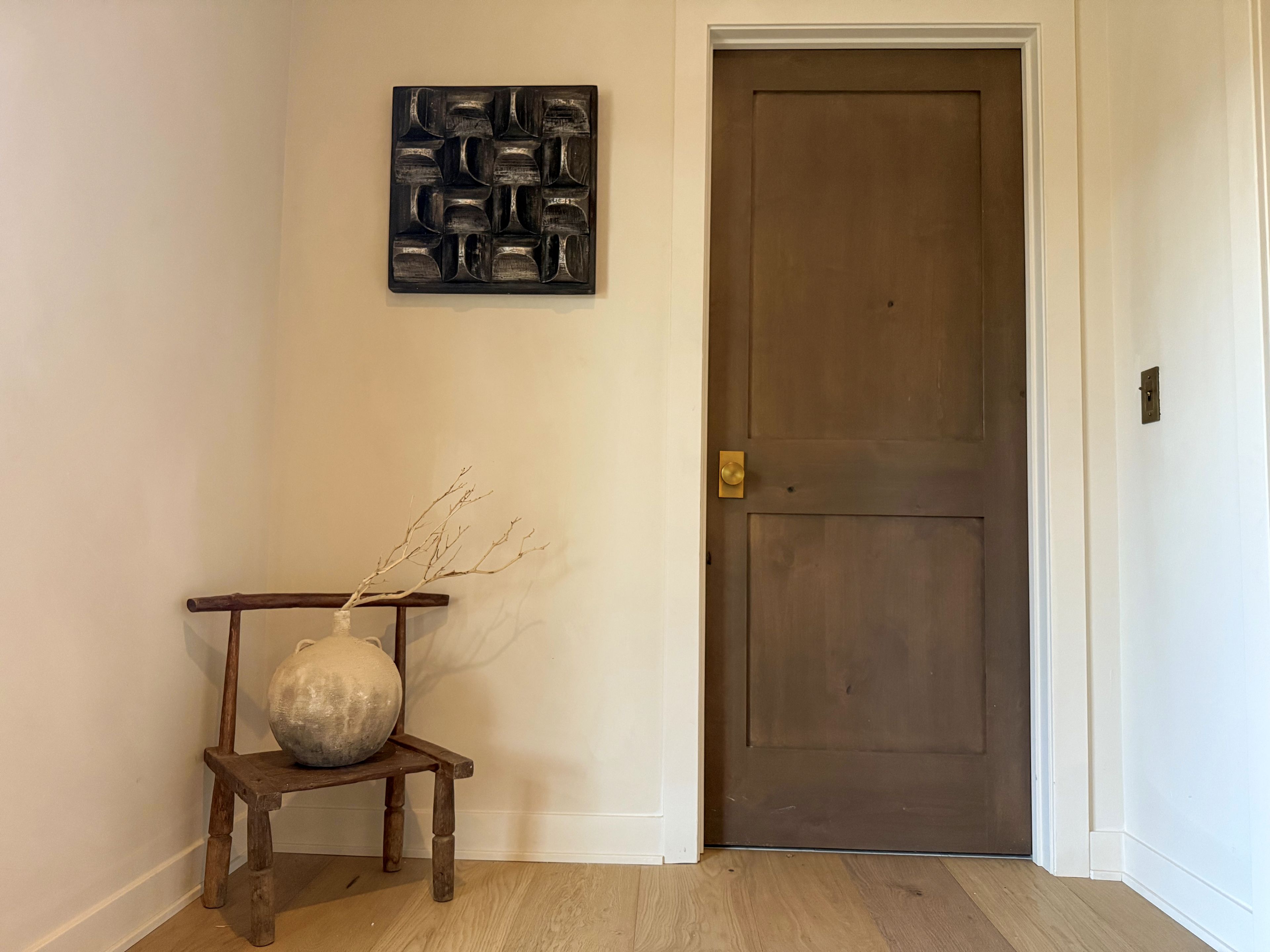 A hallway with a wooden door, a small wooden chair with a vase, and a wall-mounted abstract art piece.