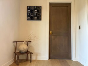 Un pasillo con una puerta de madera, una pequeña silla de madera con un jarrón y una obra de arte abstracto colgada en la pared.
