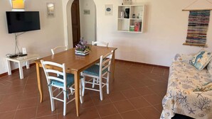 Een zithoek met een houten tafel en witte stoelen, een televisie op een dressoir, een klein plankje met spullen en een gestreept sofa.