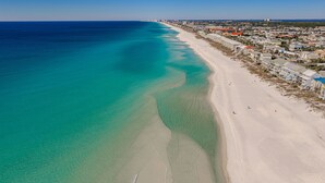 Vista aérea de una playa con aguas turquesas cristalinas, una orilla arenosa y un pueblo costero con edificios y un faro a lo lejos.