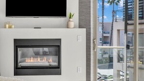 A modern living room with a fireplace, a flat-screen TV, and a balcony with a view of palm trees and buildings. - Relaxed Coastal Vibes - Luxury Accomodations & Steps to the Sand, Shops and Pier (Oceanside)