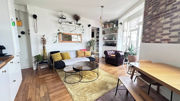 Ein modernes Wohnzimmer mit einer Couch, einem Couchtisch und einem Essbereich mit Holztisch und Stühlen. Es gibt Bücherregale, einen Kamin und ein großes Fenster mit einer Ziegelwand.
