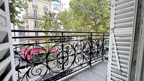 Ein Balkon mit aufwendigen schwarzen Metallgeländern, Blick auf eine Straße mit Gebäuden und einem roten Fahrzeug, das am Gehweg geparkt ist.
