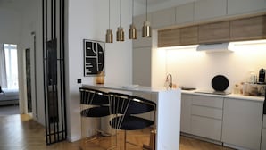 Eine moderne Küche mit weißer Arbeitsplatte, zwei schwarzen Barhocker, Pendelleuchten und einem großen Fenster.