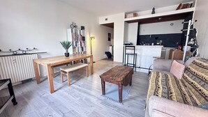 Een moderne woonkamer met een houten eettafel, een bankje en een bank. Achtergrond toont een boekenkast en een klein keukentje.