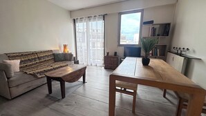 Een woonkamer met een bank, salontafel en een eettafel. Er is een boekenkast en een radiator.