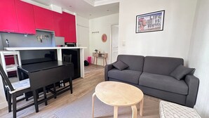 Un espace de vie compact comprenant un coin cuisine, un canapé gris, une petite table ronde et un tableau accroché au mur.