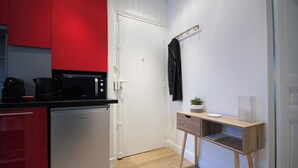 Une cuisine compacte équipée d’un four à micro-ondes, d’une machine à café et d’un lave-vaisselle. Un petit bureau en bois, agrémenté d’une plante et d’un vase, est adossé au mur.