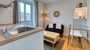 A modern kitchen with a stainless steel sink and a window with curtains. There is a black sofa with white cushions and a glass coffee table. A floor lamp and a decorative mirror are on the wall. - On Room Last Floor - Batignolles (Paris)