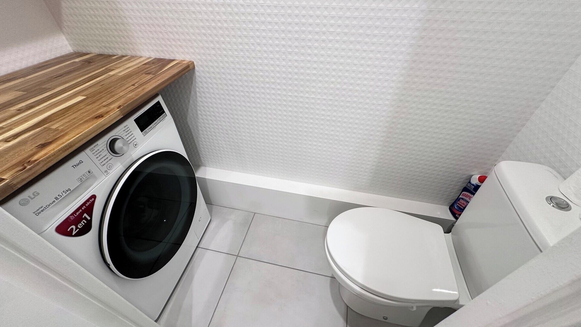 A compact bathroom with a washing machine, toilet, and a wooden shelf.