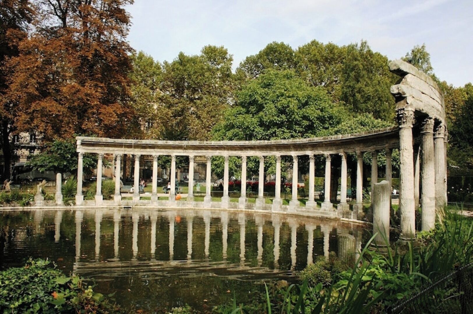 Een klassiek stenen gebouw met kolommen, een spiegelglad wateroppervlak en weelderige groene begroeiing op de achtergrond.