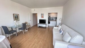 A modern living room with a glass dining table, two chairs, a sofa, a TV on a stand, and a kitchen in the background. - Studio balcony and fiber – Parc Monceau 8th (Paris)
